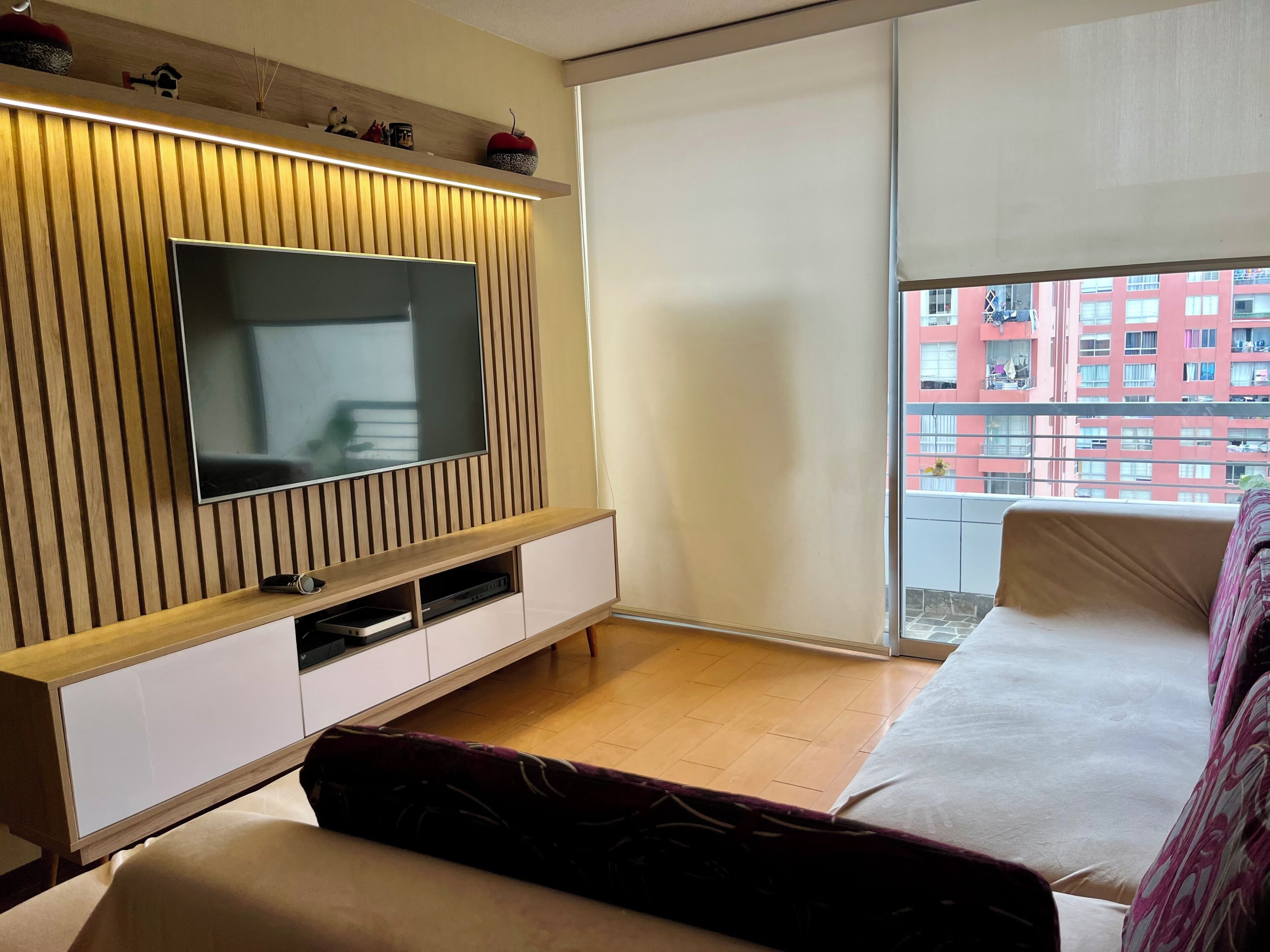 A modern living room with a flat-screen TV, wooden paneling, a sofa, and a balcony with a view of a red building.