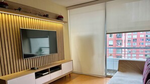 Ein modernes Wohnzimmer mit Flachbildfernseher, Holzvertäfelung, einer Couch und einem Balkon mit Blick auf ein rotes Gebäude.