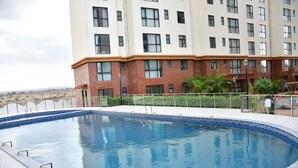 Une piscine dont le bord est recouvert de carreaux, entourée d’une clôture, et un immeuble à plusieurs étages avec une façade en briques en arrière-plan.