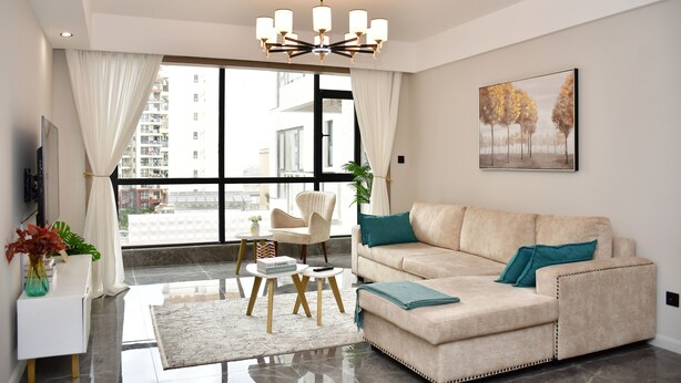Un salon moderne avec un canapé beige, un lustre, une grande fenêtre avec des rideaux, un tableau au mur et une plante sur la table d’appoint.