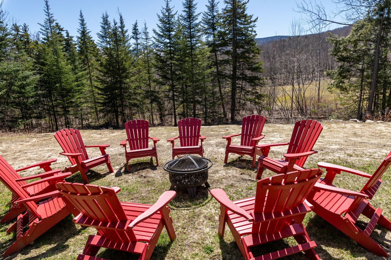 Sillones Adirondack rojos dispuestos en círculo alrededor de un brasero en un espacio al aire libre con césped y árboles de fondo.