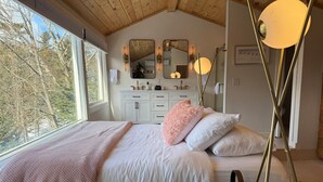 Une chambre avec une grande fenêtre, un lit recouvert de draps blancs, une table de chevet et un miroir au-dessus de la commode.