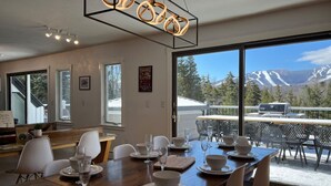 Un coin repas avec une table en bois dressée pour quatre personnes, de grandes fenêtres offrant une vue sur des montagnes enneigées et un luminaire suspendu moderne.