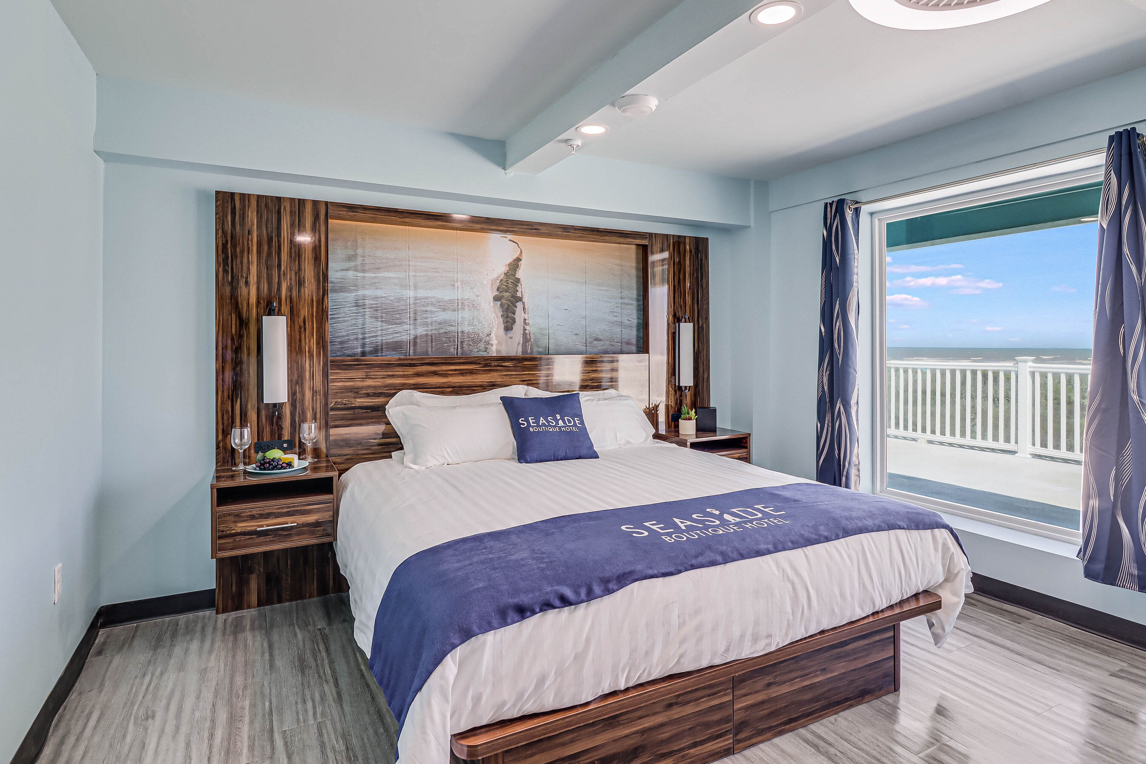 Quarto de hotel moderno com uma cama grande, cabeceira de madeira, vista para o mar e varanda com cortinas.