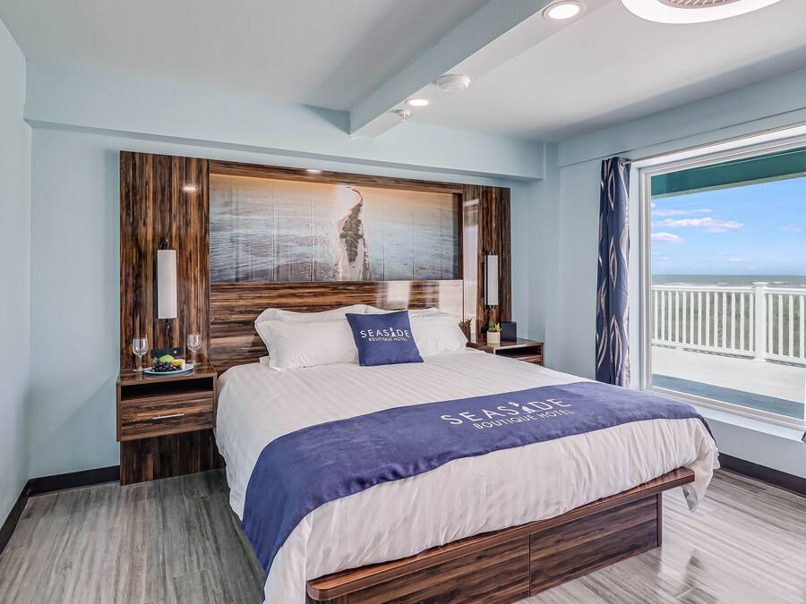 A modern hotel room with a large bed, a wooden headboard, a view of the sea, and a balcony with curtains.