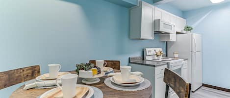 A dining area with a wooden table set for four, white plates, bowls, and cups. A microwave and refrigerator are visible in the background.