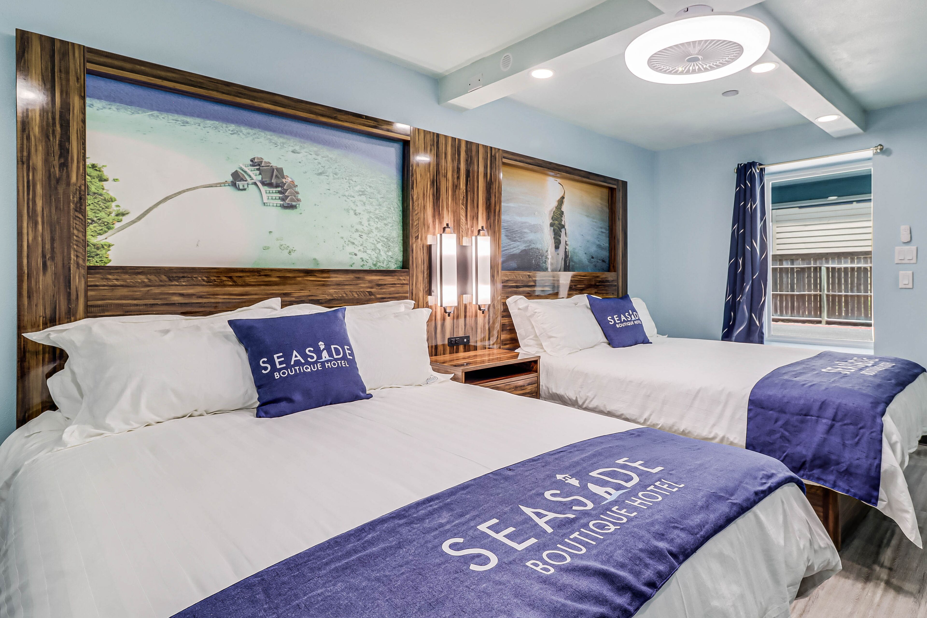 A hotel room with two beds, each with a 'Seaside Boutique Hotel' bedspread, wooden headboards, and a large framed seascape on the wall.