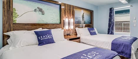 A hotel room with two beds, each with a 'Seaside Boutique Hotel' bedspread, wooden headboards, and a large framed seascape on the wall.