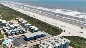 An aerial view of a coastal area with a beach, a parking lot, and several buildings.