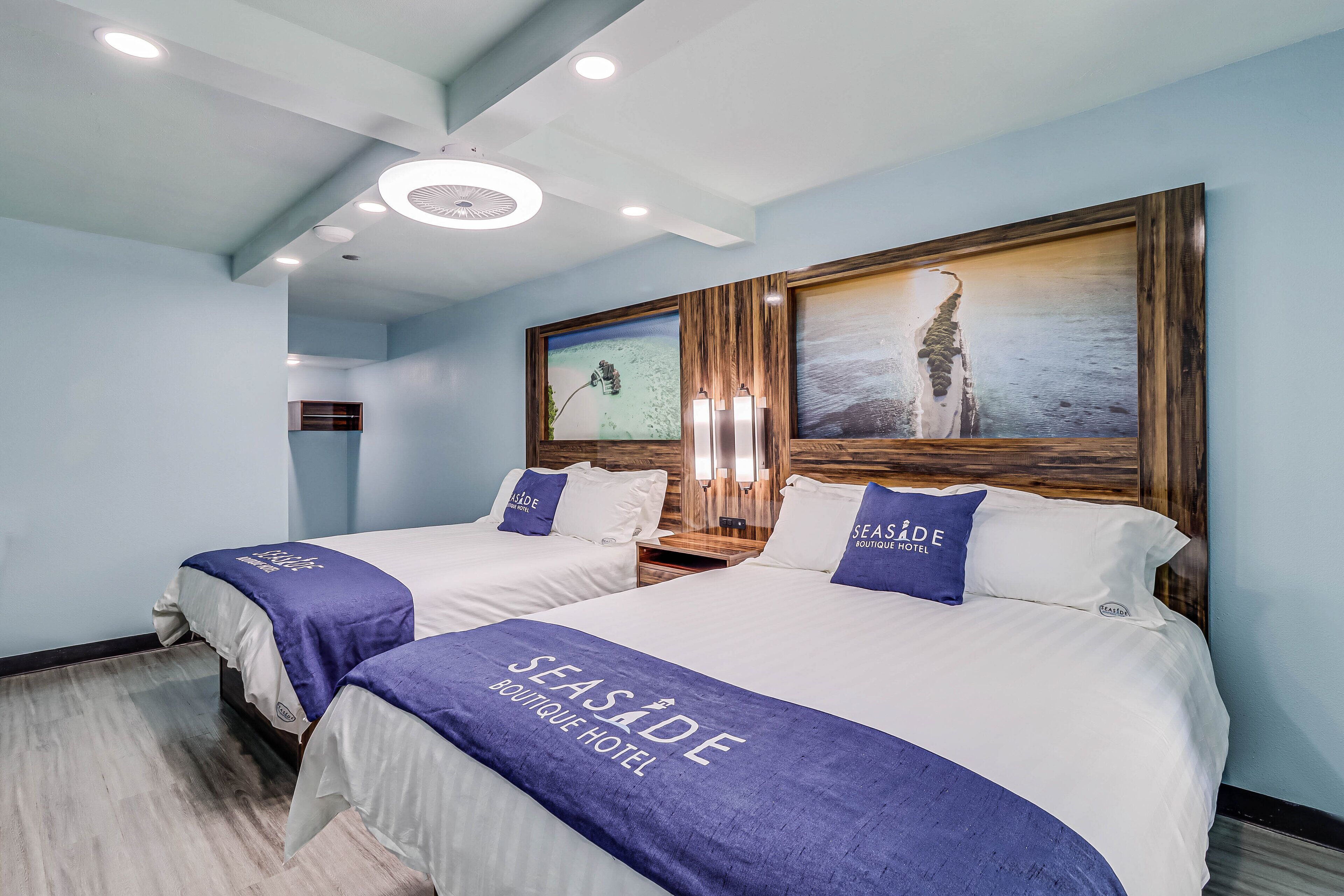 Una habitación de hotel con dos camas, cada una con una colcha del 'Seaside Boutique Hotel', una obra de arte enmarco grande sobre las camas y un cabecero de madera.