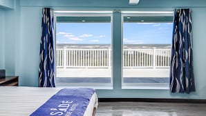 A hotel room with a large window, a bed with a blue bedspread, and a view of a white fence and sky.