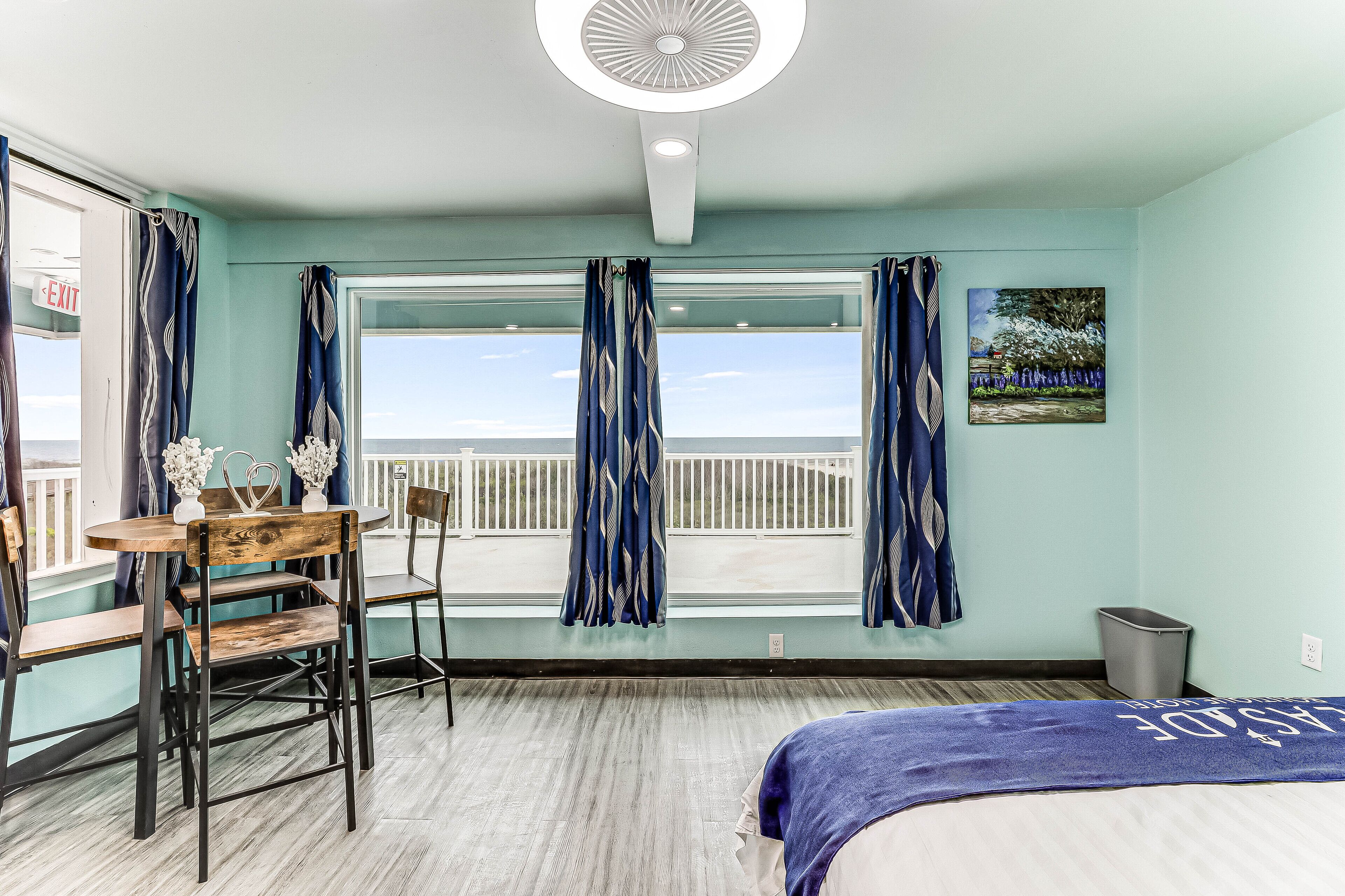 A room with a bed, a table with chairs, a window with curtains, a painting on the wall, and a balcony with a railing.