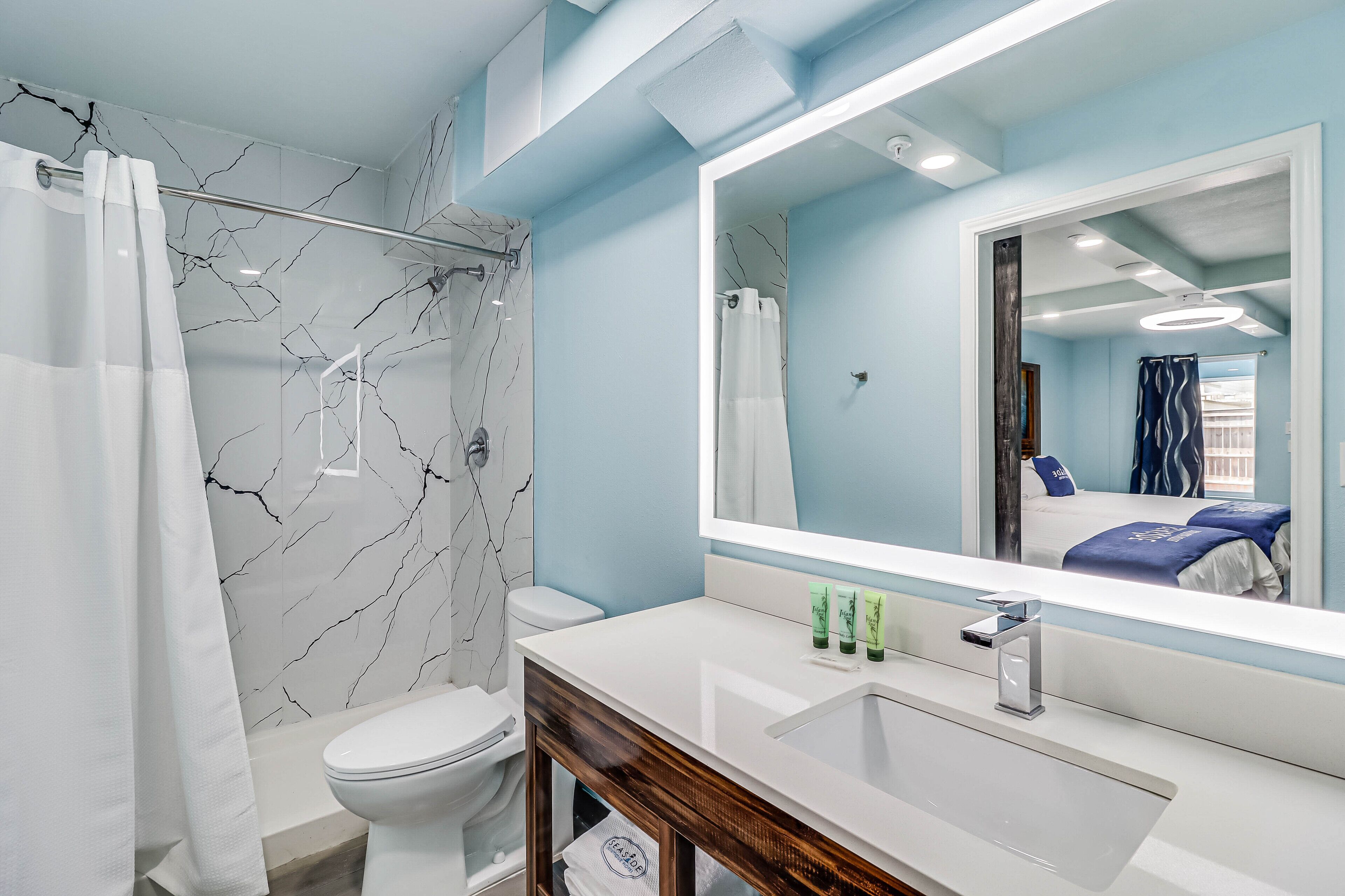 Un baño moderno con pared de ducha de mármol, inodoro blanco y un espejo grande que refleja una cama con ropa de cama azul.