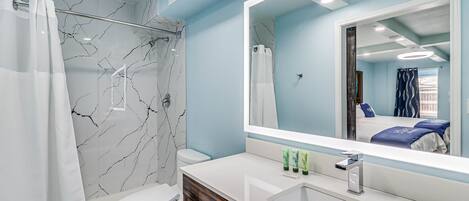 Un baño moderno con pared de ducha de mármol, inodoro blanco y un espejo grande que refleja una cama con ropa de cama azul.