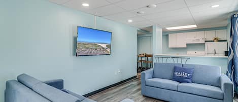 A modern hotel room with a light blue color scheme, a large flat-screen TV mounted on the wall, a sofa with a pillow, and a kitchenette area in the background.