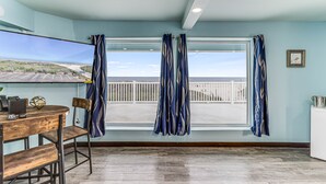 A room with a large window, a wooden table, a chair, and a view of the beach.