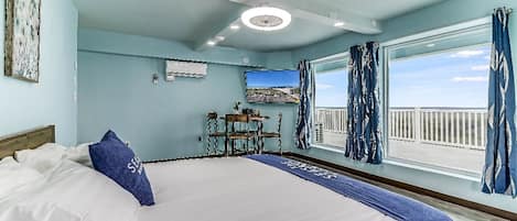 A bedroom with a large bed, a desk with chairs, a view of the sea, and a ceiling fan.
