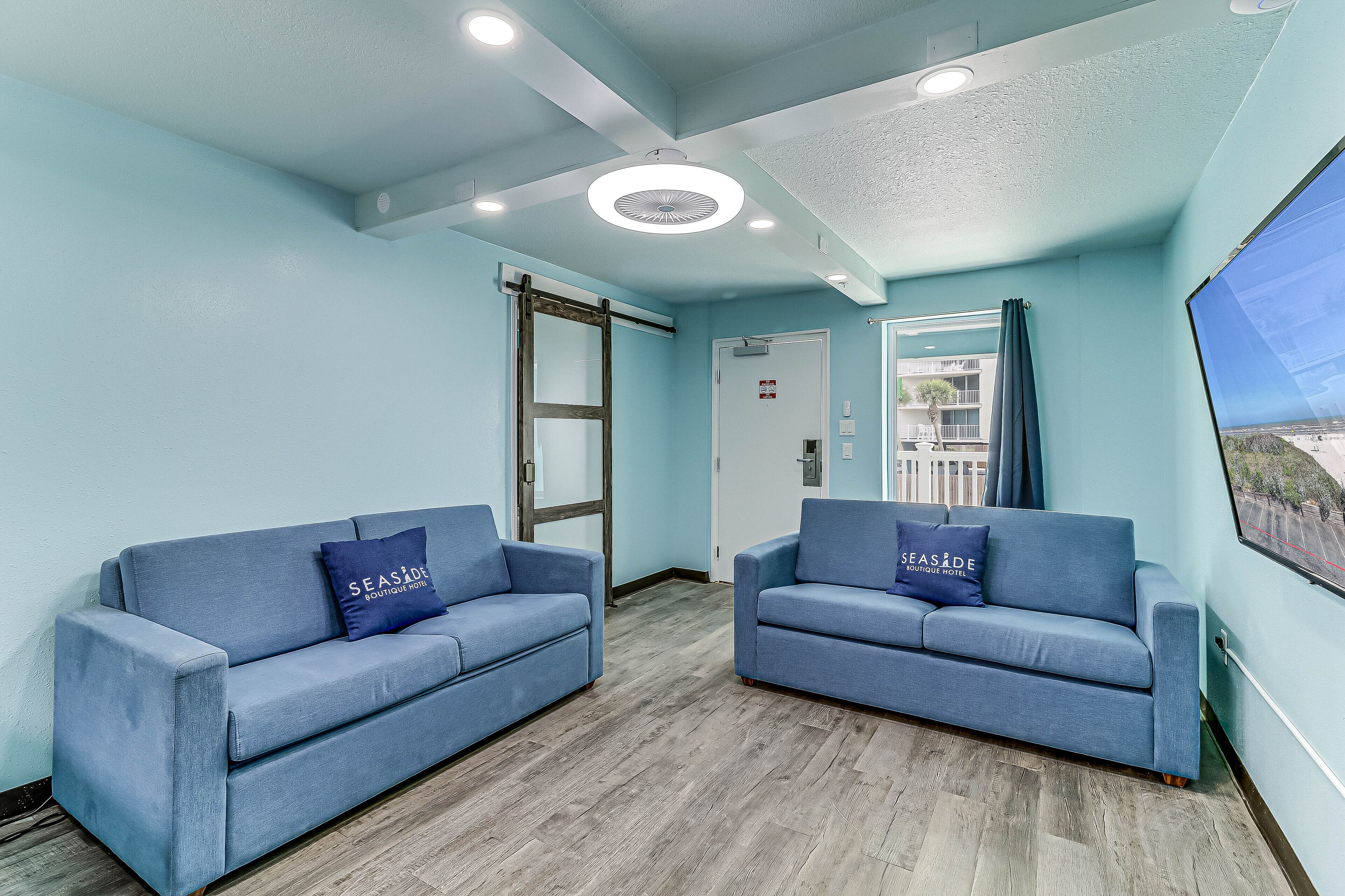 Una habitación con dos sofás azules, suelo de madera, techo con iluminación empotrada y vista por una ventana.