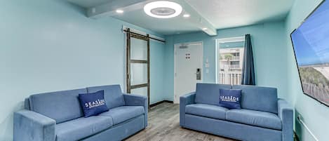 A room with two blue sofas, a wooden floor, a ceiling with recessed lighting, and a view through a window.