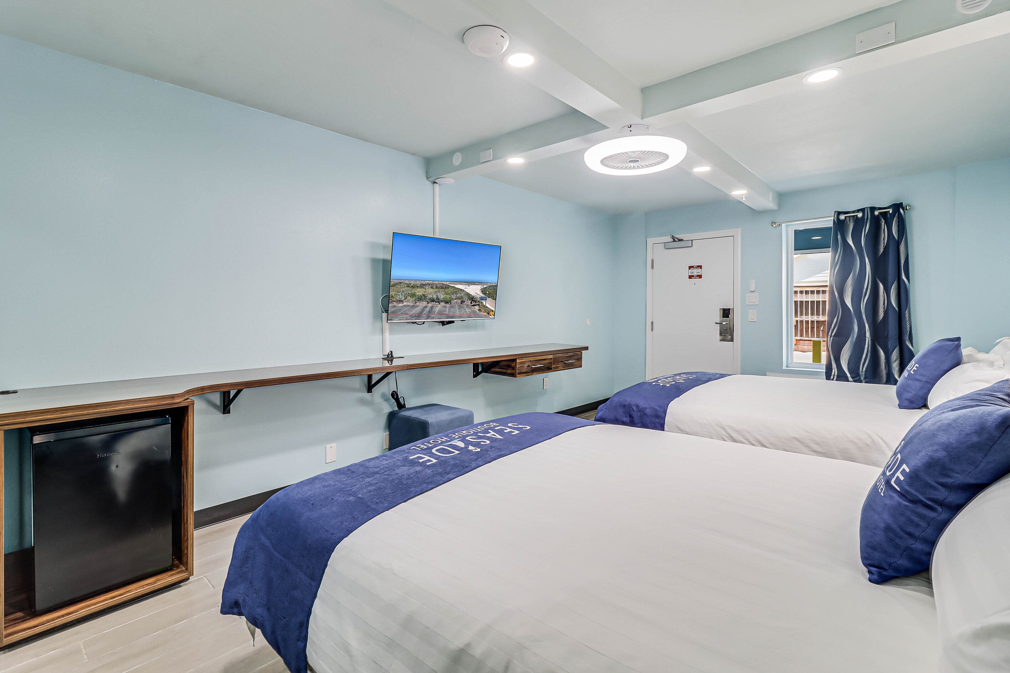 Habitació d'hotel amb dos llits, un televisor fixat a la paret, un ventilador de sostre i una finestra amb cortines.