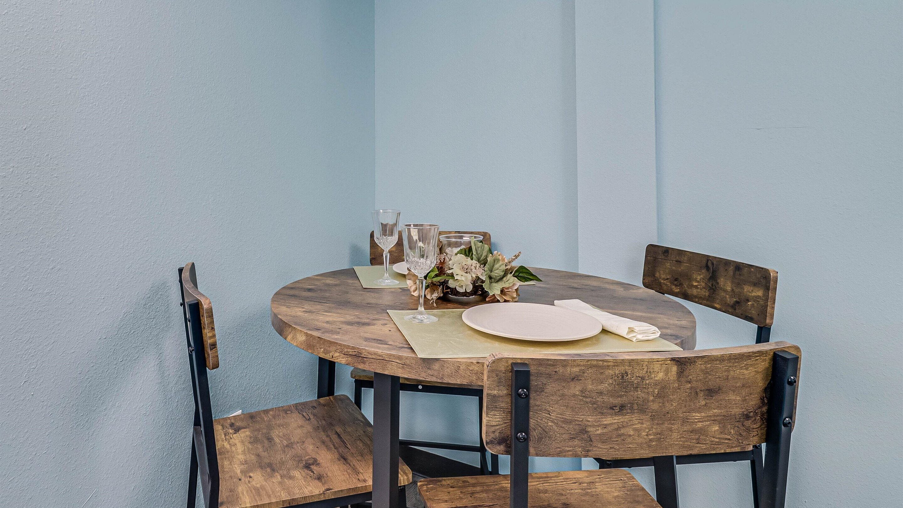 A round wooden table with a placemat, two white plates, two wine glasses, and a small floral arrangement.