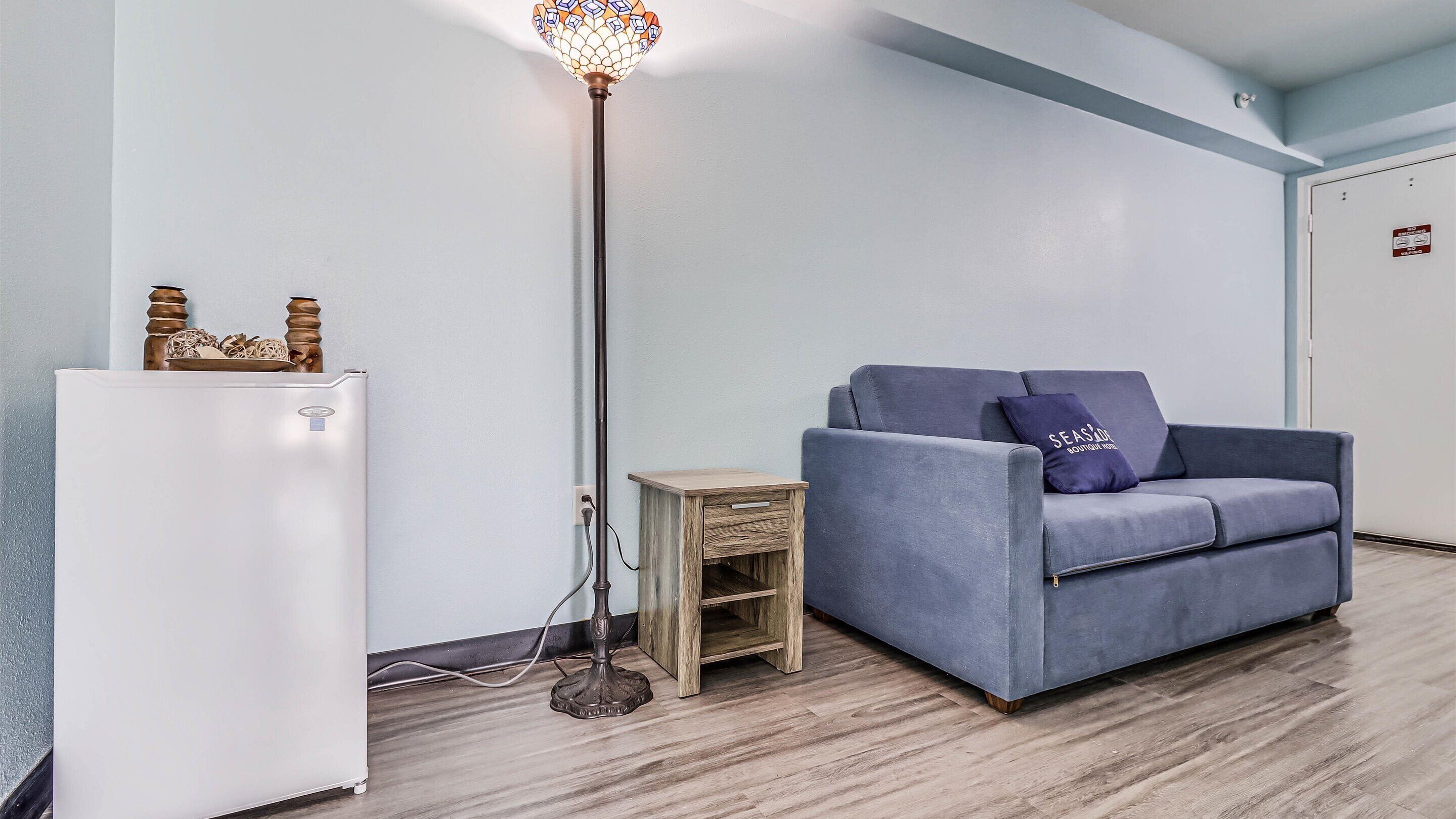 A room with a white refrigerator, a blue sofa with a cushion, a floor lamp, and a small wooden side table.