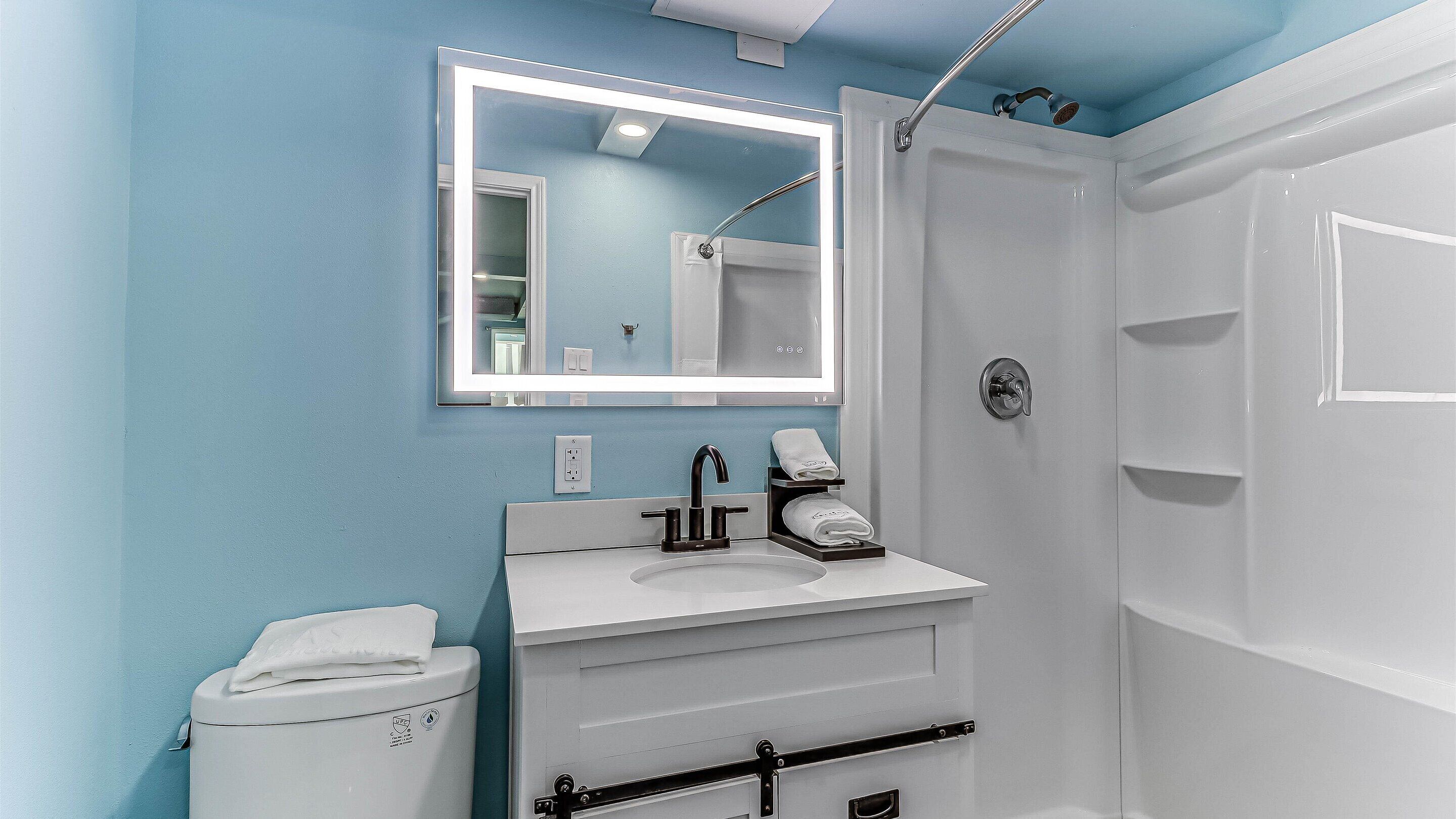 A bathroom with a white sink, a mirror with a light, a shower, and a toilet.