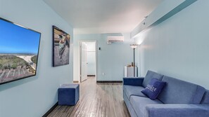 Une chambre d’hôtel moderne dotée d’une grande fenêtre donnant sur la plage, d’un canapé bleu avec un coussin, d’un petit pouf et d’un lampadaire.
