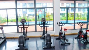 Et fitnessrum med tre motionscykler, et løbebånd og en ro-maskine, store vinduer giver udsigt til en swimmingpool og grøn bevoksning.