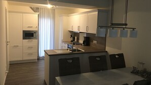 Cocina compacta con armarios blancos, microondas integrado y cafetera. Hay una zona de comedor con mesa y sillas.