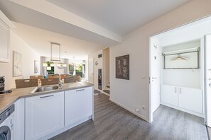 A modern kitchen with a central island, a dining area with wicker furniture, and a hallway leading to a bathroom. - Ferienhaus Nordsee-koje mit Meerblick, Strandkorb, Kamin, Sauna (Wangerland)