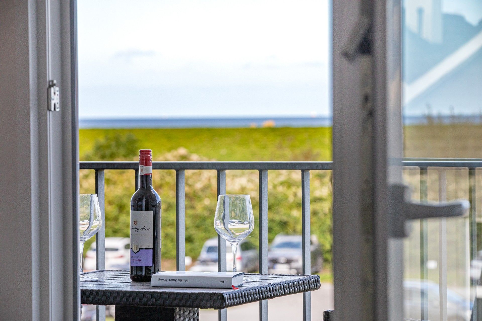 Un balcon avec une bouteille de vin et deux verres sur une table, offrant une vue sur la mer et la verdure.