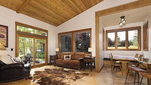 A spacious living room with a large wooden ceiling, a leather sofa, a cowhide rug, and a dining area with wooden chairs and a table. - Lone Moose Lookout | Big Sky Meadow (Big Sky)