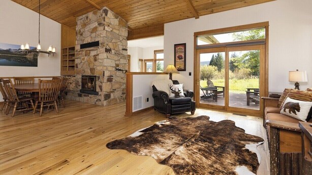 A spacious living room with a stone fireplace, wooden ceiling, and large windows offering a view of the outdoors. - Lone Moose Lookout | Big Sky Meadow (Big Sky)
