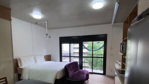 A compact hotel room with a bed, a small table, a purple chair, a kitchenette, and a balcony with a view. - Tela de Cinema! Novinho Prox. Paulista c/ Vaga (Consolação São Paulo)