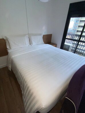 Habitación de hotel con una cama grande, una mesita de noche y una ventana con vistas a edificios.