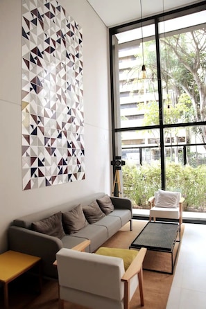 Uma sala de estar moderna com um sofá cinzento, uma poltrona branca e uma mesa pequena. Há uma janela grande com vista para áreas verdes e edifícios.
