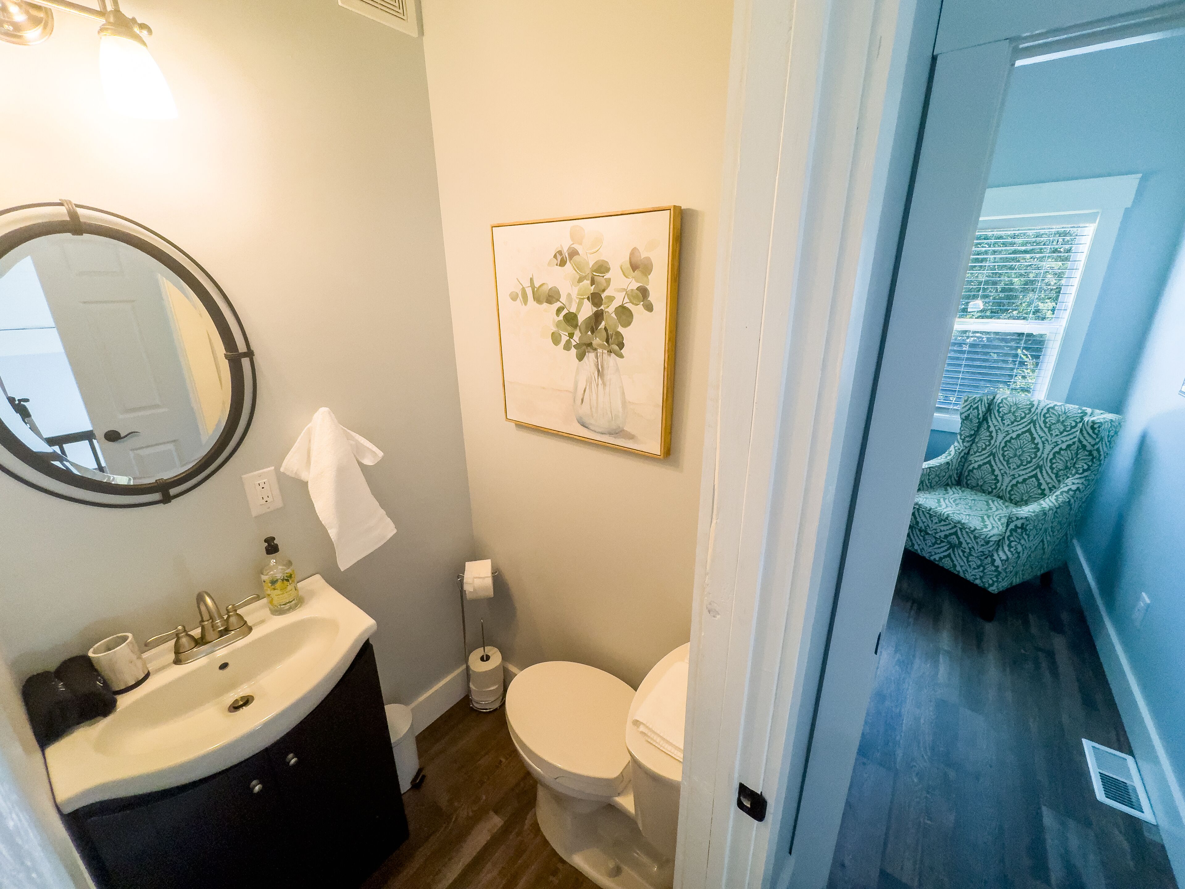 A bathroom with a round mirror, white sink, toilet, and a framed picture on the wall.