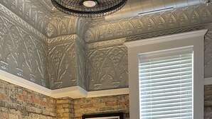 Une chambre avec un mur en briques, une affiche encadrée, une fenêtre avec des stores, un lit avec une literie à motifs et une lampe.