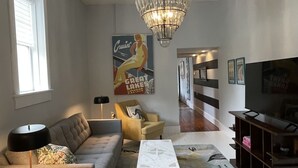 Un salon comprenant un canapé, un fauteuil, une table basse et un tapis à motifs. On y trouve également un lustre, une affiche accrochée au mur et un téléviseur posé sur un meuble.