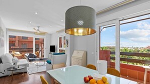 Una sala de estar moderna con una mesa de comedor de vidrio, un ventilador de techo y un balcón con vistas a zonas verdes.