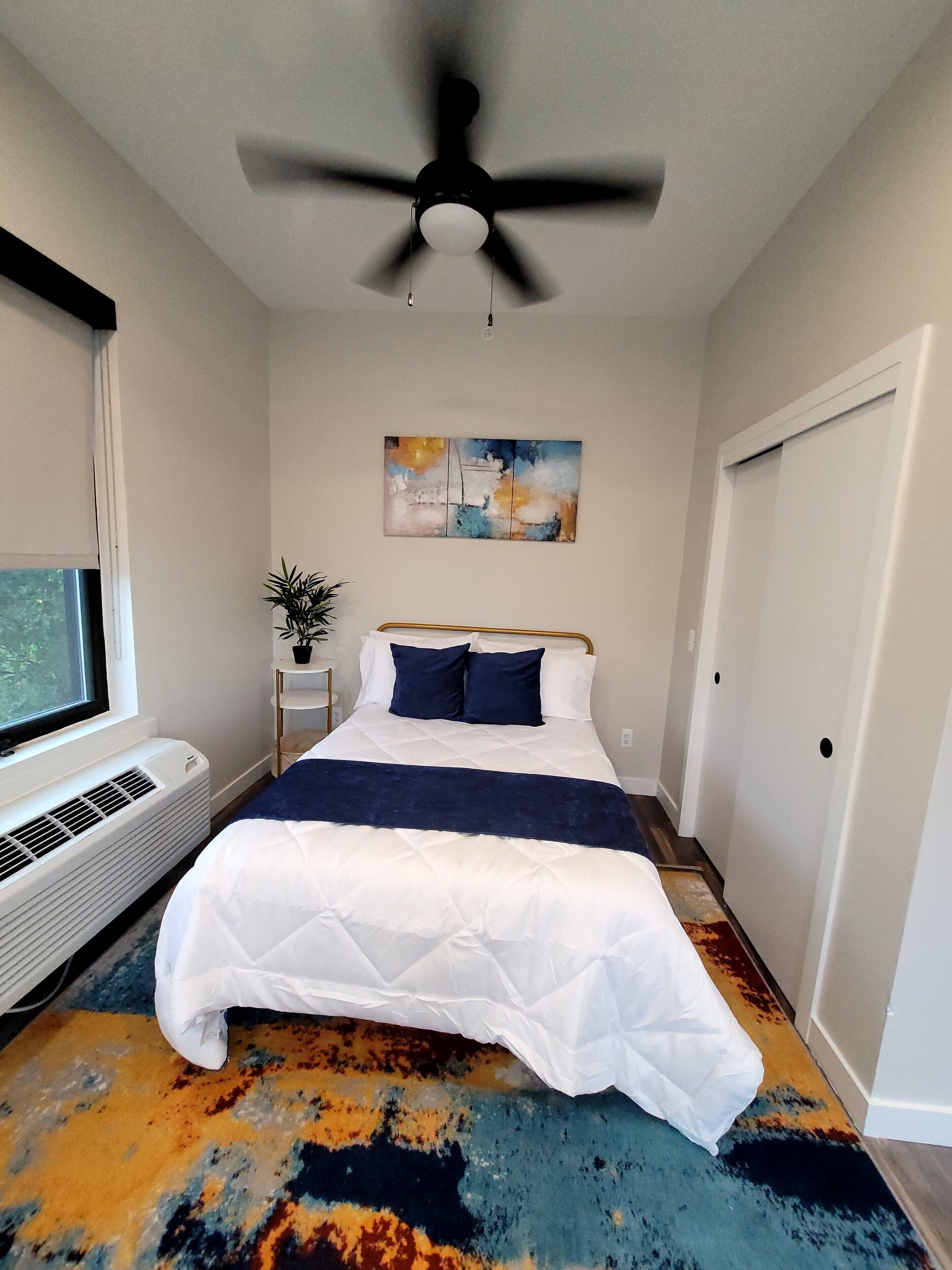 A bedroom with a bed, a ceiling fan, a painting, a heater, and a window.