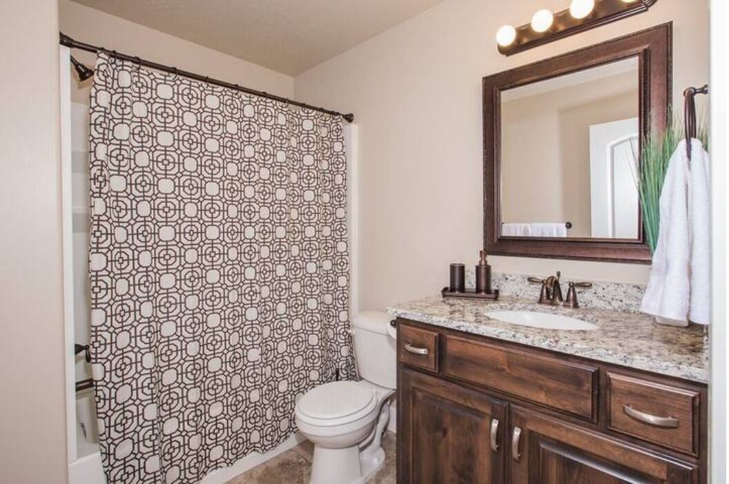 Un baño con cortina de ducha estampada, inodoro blanco, lavamanos con encimera de mármol, armarios de madera y espejo con luces arriba.