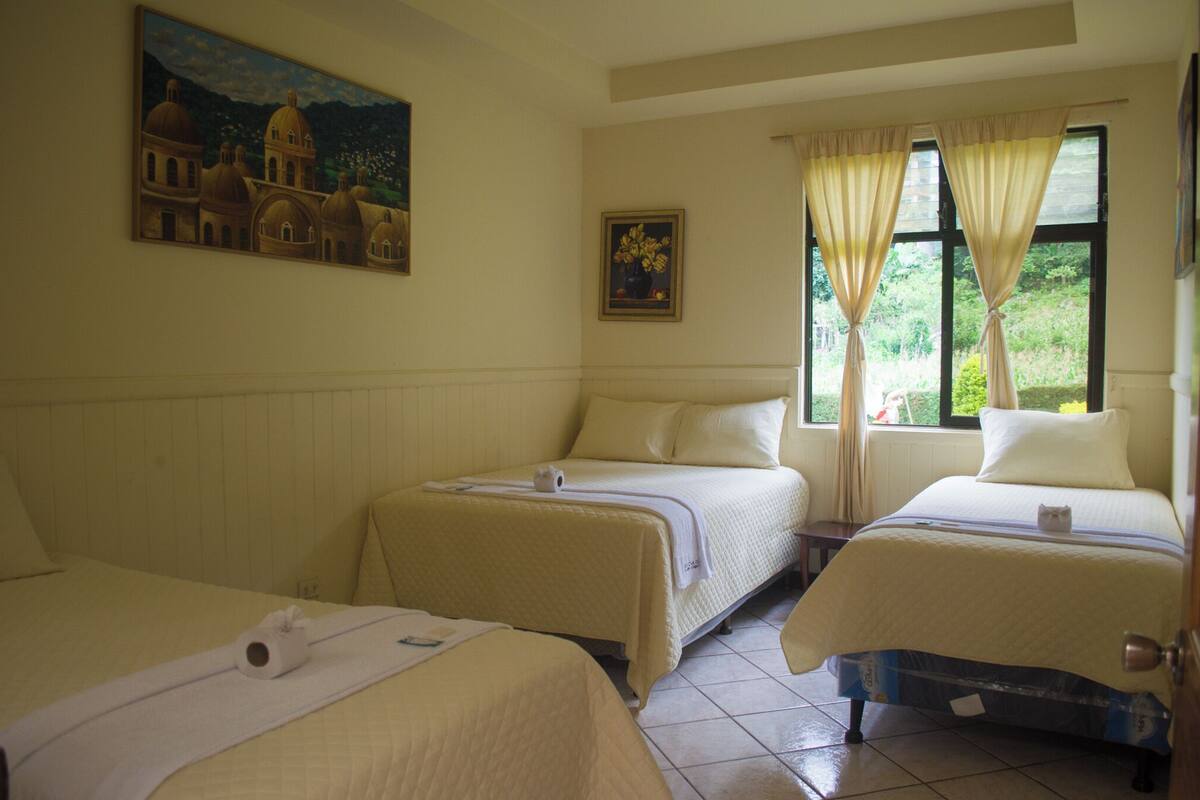 A hotel room with two beds, a painting of a building, and a window with curtains.