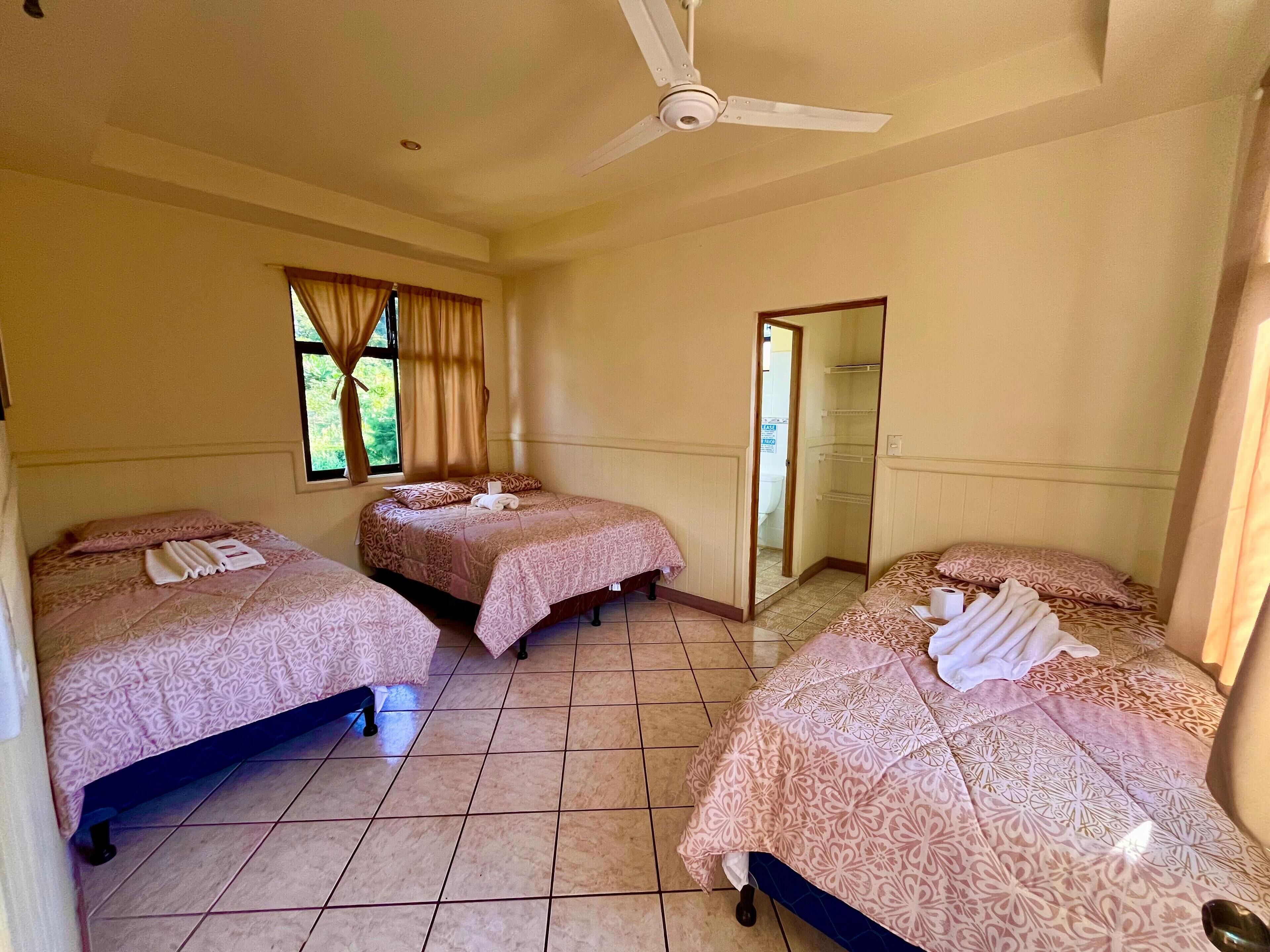 Une chambre d’hôtel avec trois lits, chacun recouvert d’une couvre-lit à motifs et agrémenté de serviettes. On y trouve un ventilateur de plafond, une fenêtre avec des rideaux et une porte qui donne sur une autre pièce.
