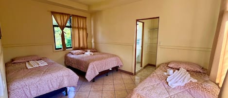 A hotel room with three beds, each with a patterned bedspread and towels. There is a ceiling fan, a window with curtains, and a door leading to another room.