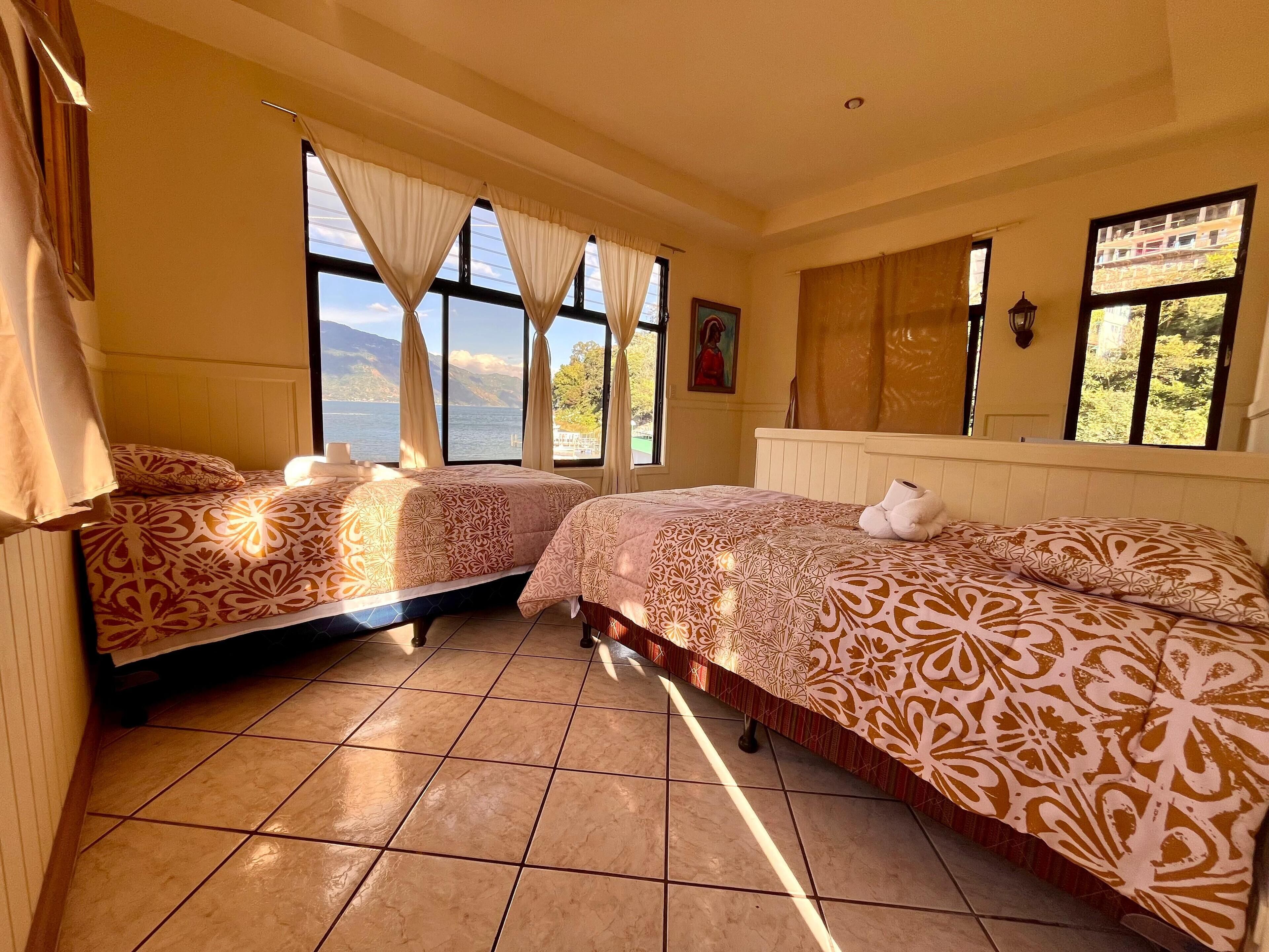 Quarto com duas camas, janelas amplas, um quadro e vista para o exterior.