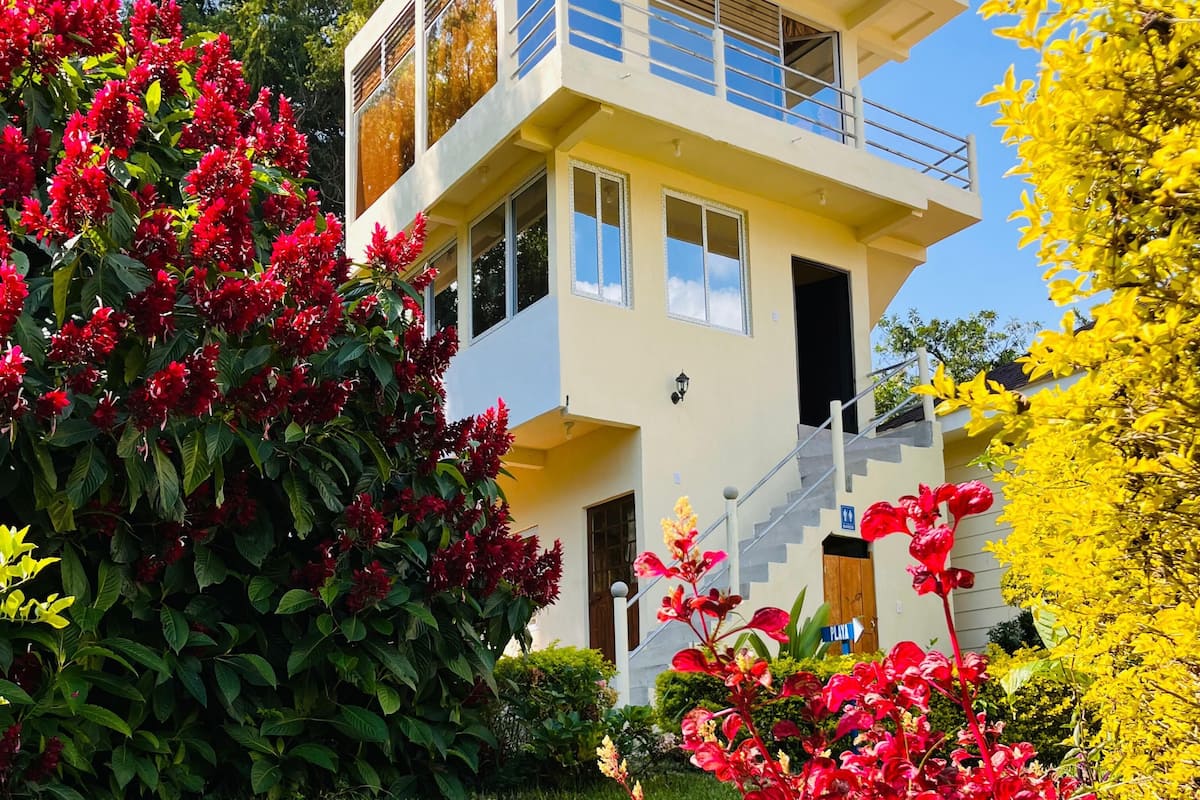 A modern two-story building with a balcony, surrounded by lush greenery and vibrant flowers.
