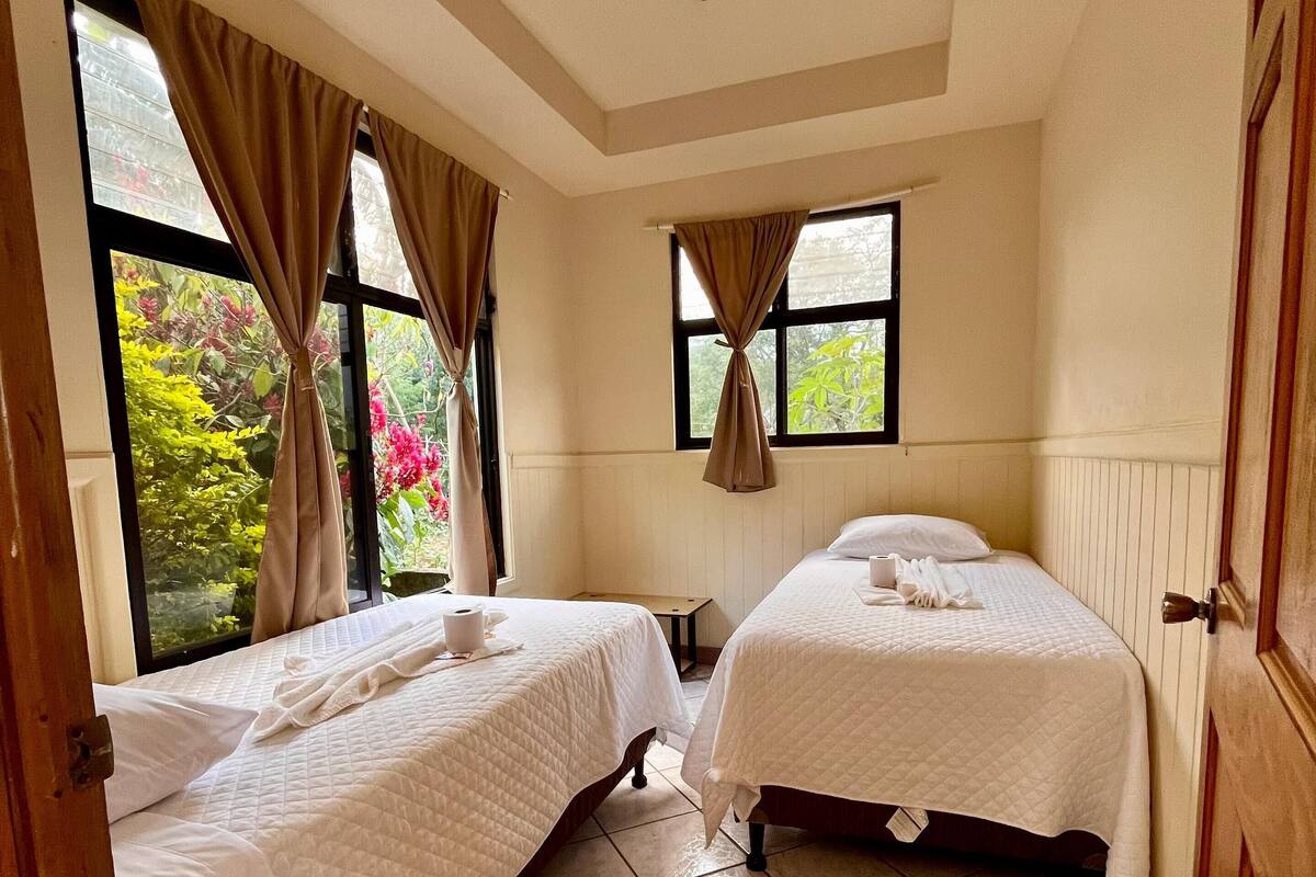 A room with two beds, a window with curtains, and a view of greenery outside.