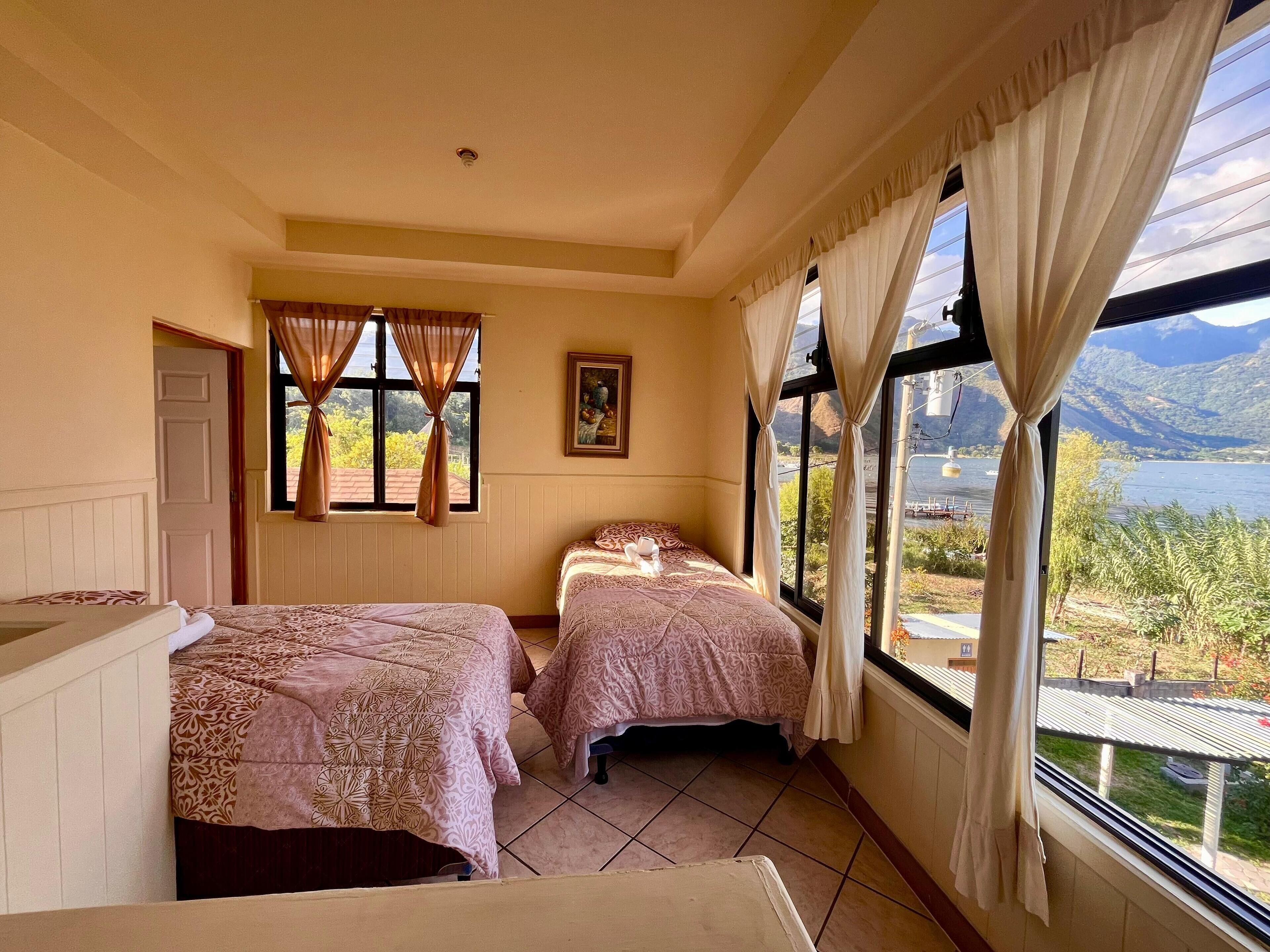 A bedroom with two beds, a window with curtains, a painting on the wall, and a view of mountains and water.